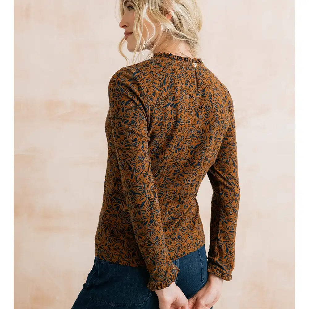 A woman shown from the back wearing a long-sleeve brown top with an intricate dark blue and orange abstract leaf pattern, paired with dark blue jeans, against a soft beige background.