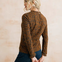 A woman shown from the back wearing a long-sleeve brown top with an intricate dark blue and orange abstract pattern, paired with dark blue jeans, set against a soft beige textured background.