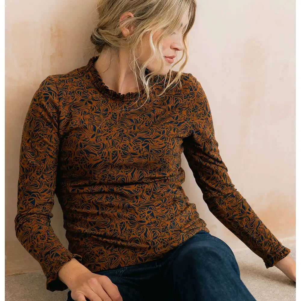 A woman wearing a long-sleeve crewneck top with a brown and black abstract leaf pattern, paired with dark blue jeans, sitting against a textured beige wall.