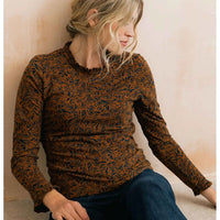 A woman wearing a long-sleeve crewneck top with a brown and black abstract leaf pattern, paired with dark blue jeans, sitting against a textured beige wall.