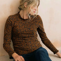 A woman wearing a long-sleeve crewneck top with a brown and black abstract leaf pattern, paired with dark blue jeans, sitting against a textured beige wall.