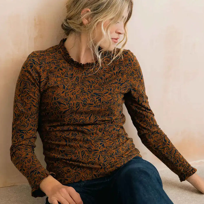 A woman wearing a long-sleeve crewneck top with a brown and black abstract leaf pattern, paired with dark blue jeans, sitting against a textured beige wall.