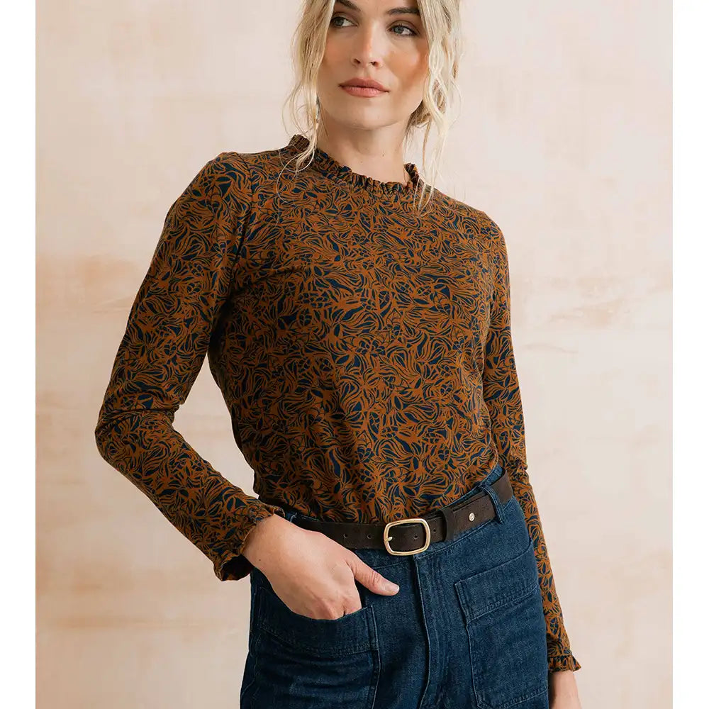 A woman wearing a long-sleeve brown and black floral patterned blouse tucked into dark blue jeans with a black belt, standing against a warm beige textured wall.