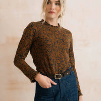 A woman wearing a long-sleeve ruffled-neck blouse with an intricate brown and black floral pattern, tucked into dark blue jeans with a black belt, set against a warm beige textured wall.