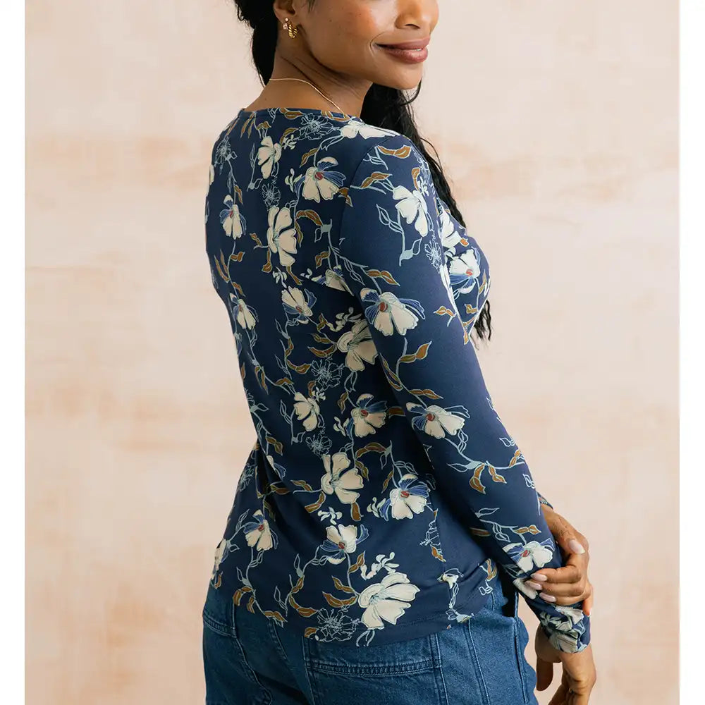 A woman wearing a navy blue long-sleeve top with a white and beige floral pattern, shown from a side-back angle against a soft beige background.