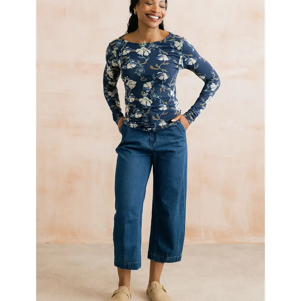 A woman modeling a navy blue long-sleeve top with a white and beige floral pattern, paired with dark blue cropped jeans and beige slip-on shoes, standing against a textured beige wall.