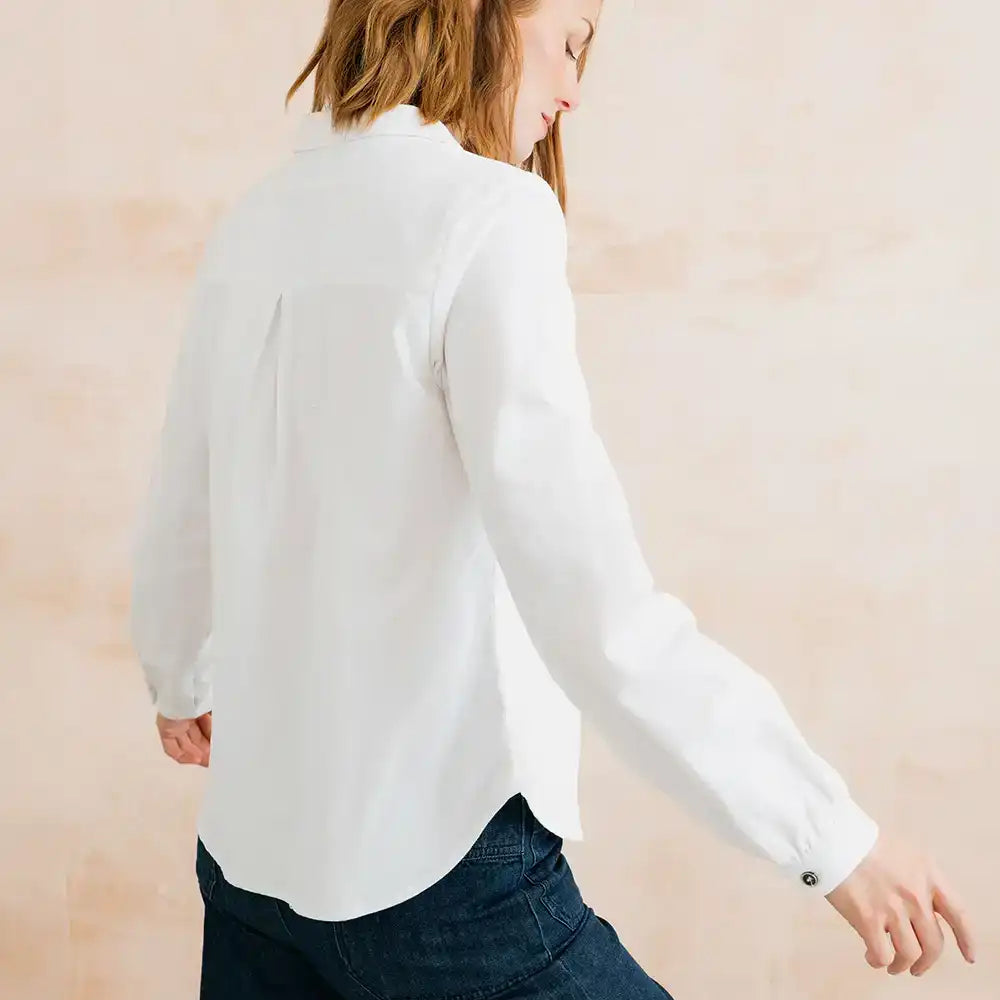 A woman wearing a white long-sleeve button-up blouse and dark blue jeans, shown from the back and side against a soft beige background.