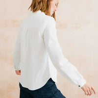 A woman wearing a white long-sleeve button-up blouse and dark blue jeans, shown from the back and side against a soft beige background.