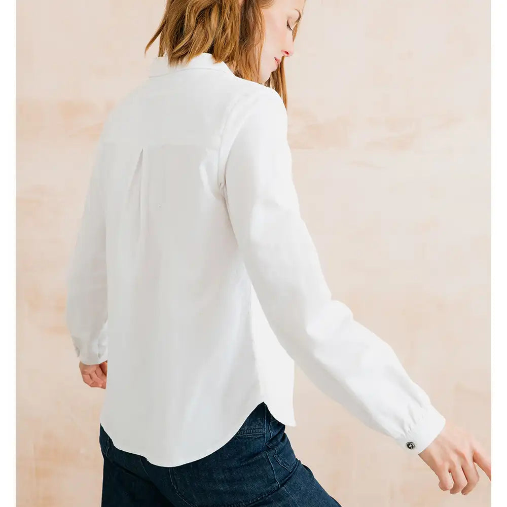 A woman wearing a white long-sleeve button-up blouse and dark blue jeans, shown from a side-back angle against a soft beige textured background.