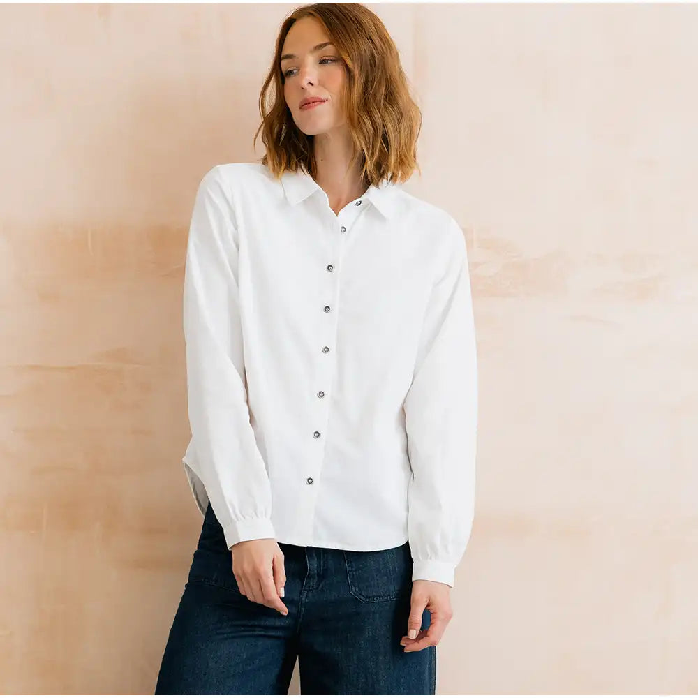 A woman wearing a white long-sleeve button-up shirt and dark blue jeans, standing against a soft beige textured wall.