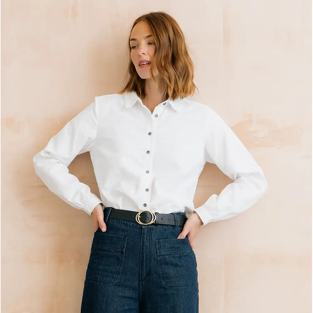 A woman wearing a white long-sleeve button-down shirt tucked into dark blue jeans with a black belt, standing against a light beige textured wall.
