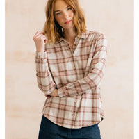 A woman wearing a long-sleeve button-up plaid shirt in cream, beige, and muted red tones, paired with dark blue jeans, standing against a light beige textured background.