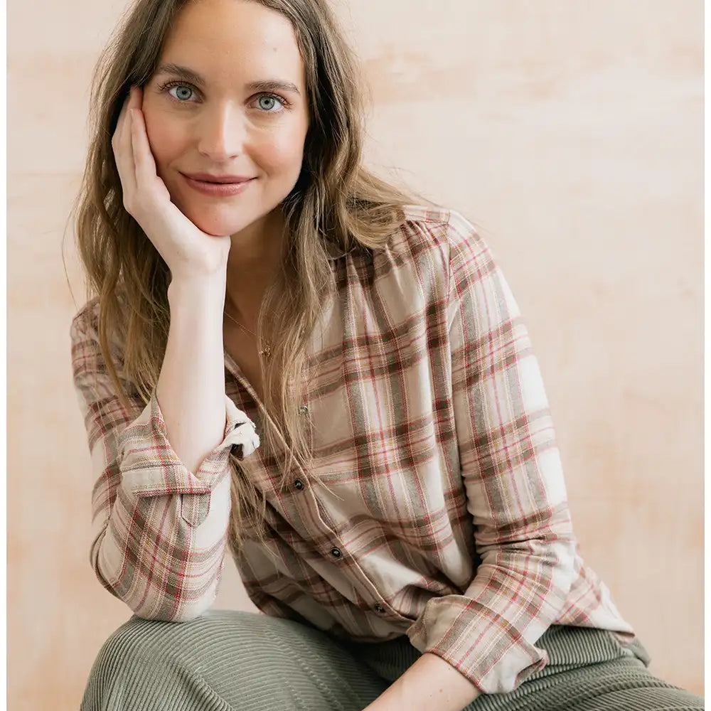 A woman with long light brown hair sits in a warm, softly lit setting, wearing a beige and brown plaid button-up shirt and olive green corduroy pants.