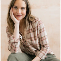 A woman with long light brown hair sits in a warm, softly lit setting, wearing a beige and brown plaid button-up shirt and olive green corduroy pants.