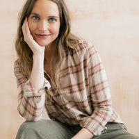 A woman with long light brown hair rests her head on her hand, wearing a beige and brown plaid button-down shirt and olive green corduroy pants, set against a soft beige background.