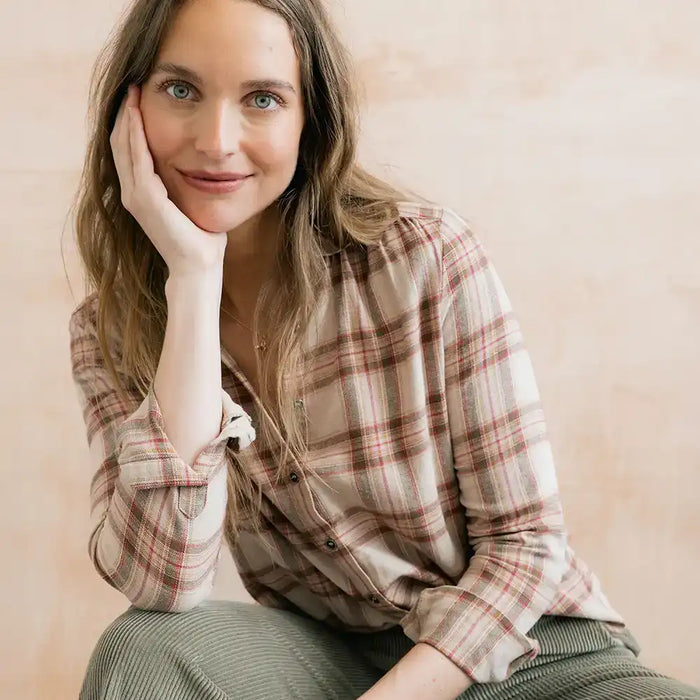 A woman with long light brown hair rests her head on her hand, wearing a beige and brown plaid button-down shirt and olive green corduroy pants, set against a soft beige background.