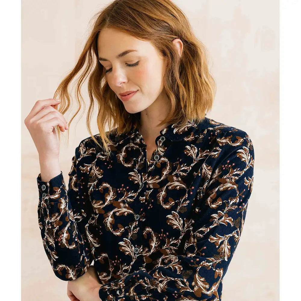 A woman wearing a long-sleeve button-up blouse with a dark navy base and an intricate gold and beige baroque-style floral pattern, set against a soft, light beige background.