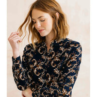 A woman wearing a long-sleeve button-up blouse with a dark navy base and an intricate gold and beige baroque-style floral pattern, set against a soft, light beige background.