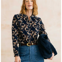 A woman wearing a navy long-sleeve button-up shirt with a beige and white floral pattern, tucked into blue jeans with a brown belt, while holding a dark navy corduroy jacket against a light beige background.