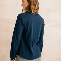 A woman shown from the back wearing a dark navy blue long-sleeve button-up shirt against a light beige textured wall.