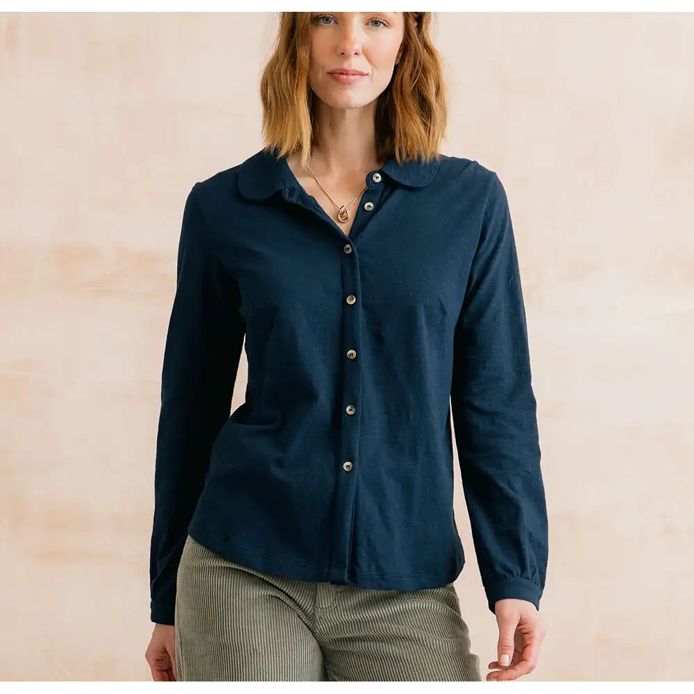 A woman wearing a dark navy blue long-sleeve button-up shirt with a collar, paired with olive green corduroy pants, standing against a light beige textured wall.