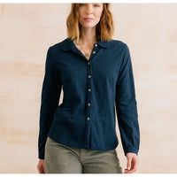 A woman wearing a dark navy blue long-sleeve button-up shirt with a collar, paired with olive green corduroy pants, standing against a light beige textured wall.