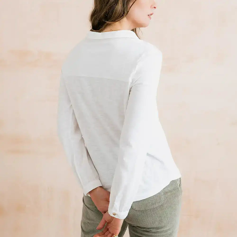 A woman wearing a white long-sleeve collared shirt with a contrasting light gray color-block panel across the upper back, shown from a side-back angle against a soft beige background.