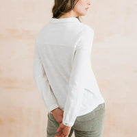 A woman wearing a white long-sleeve collared shirt with a contrasting light gray color-block panel across the upper back, shown from a side-back angle against a soft beige background.