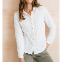 A woman wearing a white long-sleeve button-up linen shirt with gold buttons, paired with olive green corduroy pants, standing against a warm beige textured wall.