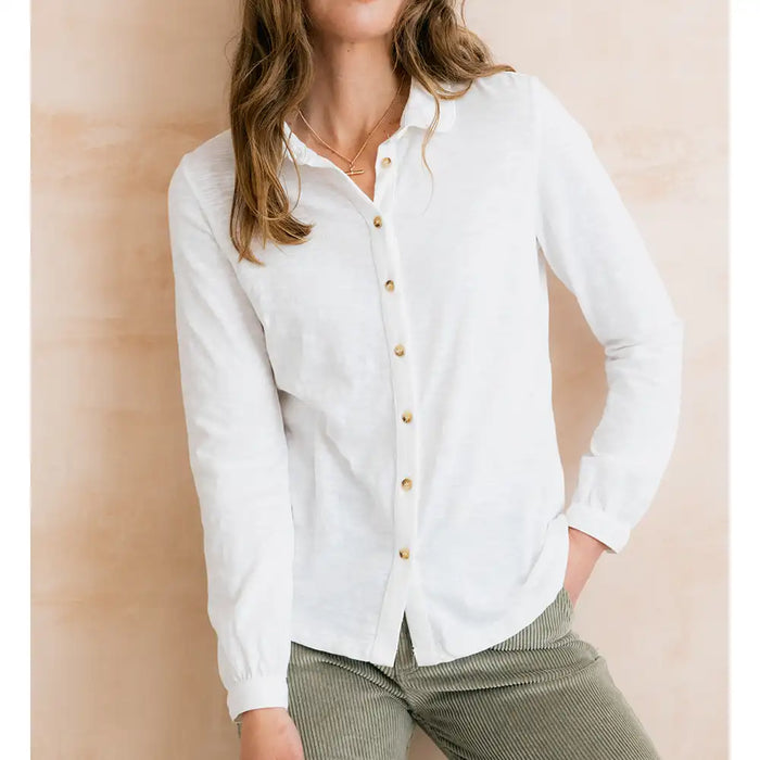 A woman wearing a white long-sleeve button-up linen shirt with gold buttons, paired with olive green corduroy pants, standing against a warm beige textured wall.