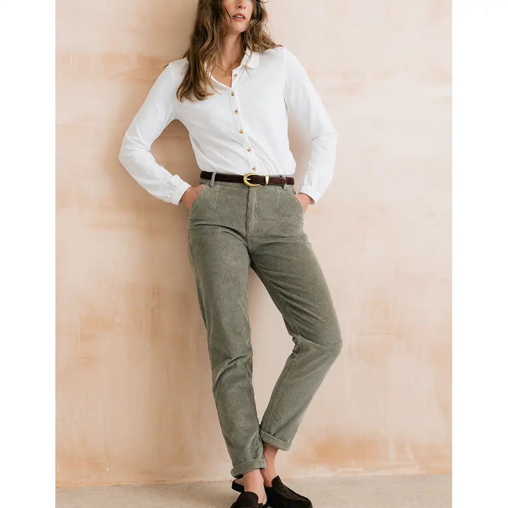 A woman models a pair of relaxed-fit olive green corduroy pants, styled with a white button-down shirt, a brown belt, and black slip-on shoes, standing against a textured beige wall.