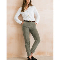 A woman models a pair of relaxed-fit olive green corduroy pants, styled with a white button-down shirt, a brown belt, and black slip-on shoes, standing against a textured beige wall.