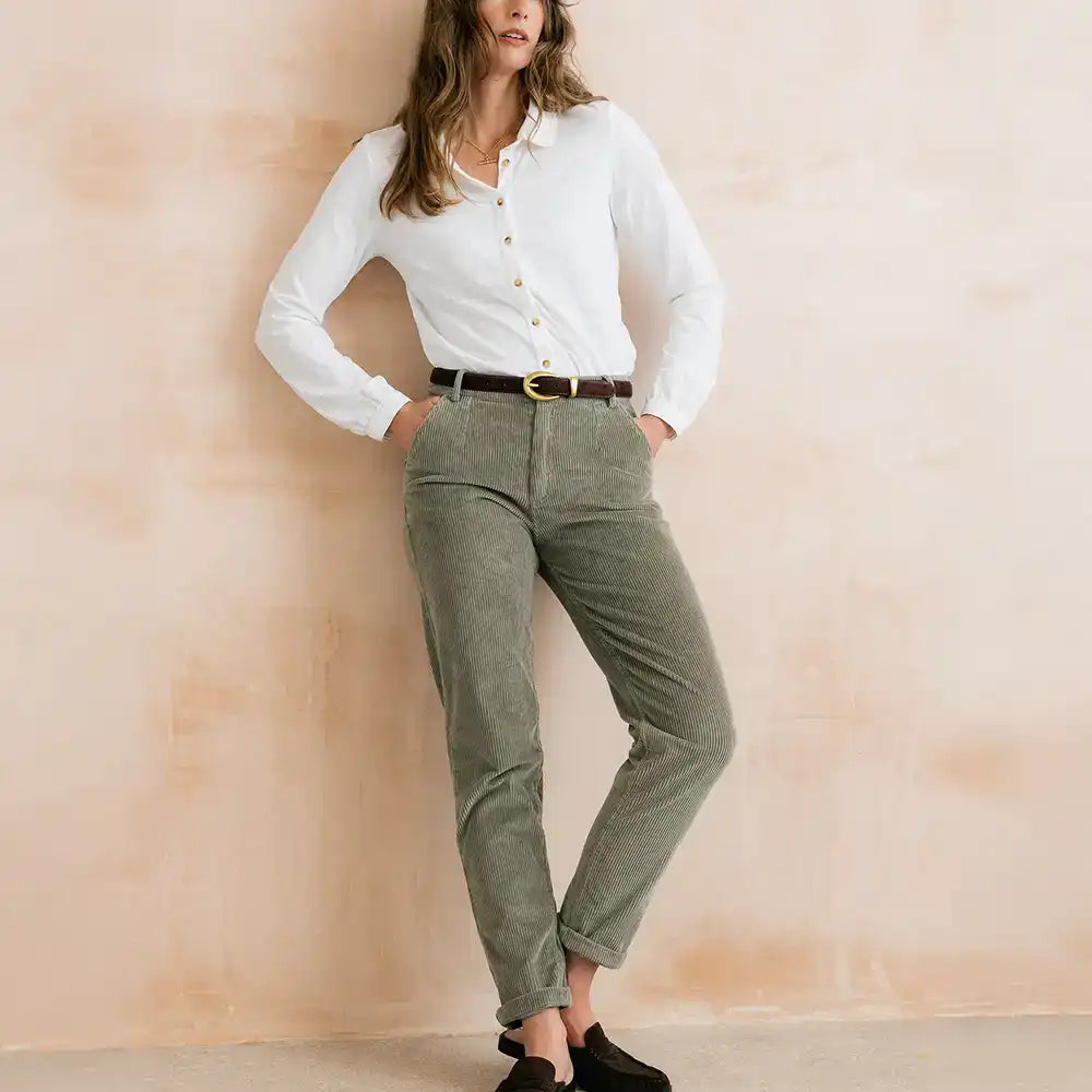 A woman models a pair of olive green corduroy trousers with a brown belt, paired with a white button-down shirt and black slip-on shoes, standing against a textured beige wall.