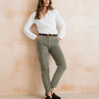 A woman models a pair of olive green corduroy trousers with a brown belt, paired with a white button-down shirt and black slip-on shoes, standing against a textured beige wall.