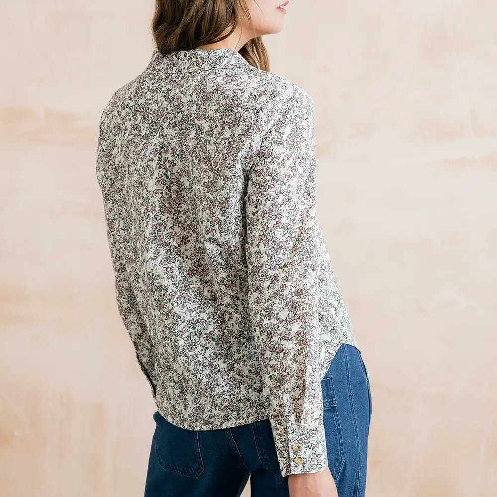 A woman shown from the back wearing a long-sleeve button-up blouse with a dense small floral print in muted brown, beige, and white tones, paired with blue jeans against a soft beige wall.