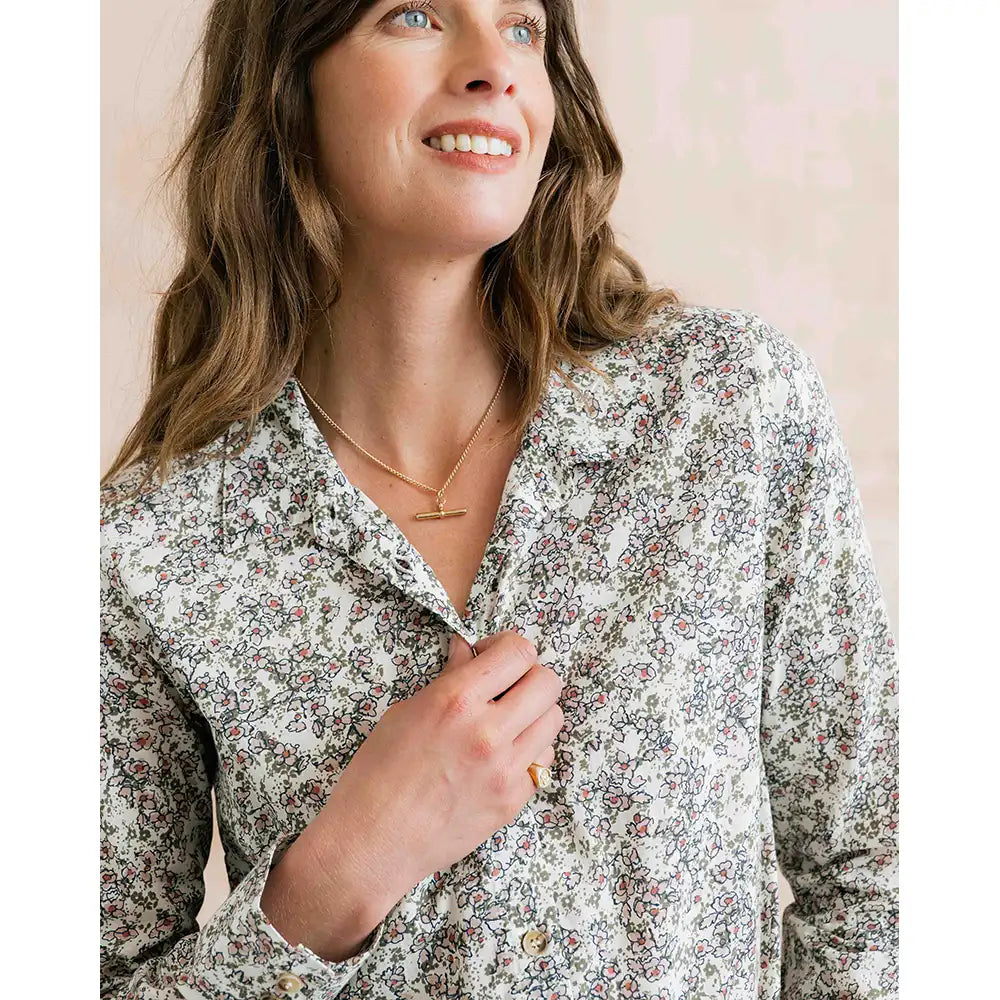 A woman wearing a long-sleeve button-up shirt with a dense multicolor floral and paisley print on a light cream background. She is smiling and looking upward, wearing a gold necklace.