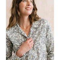 A woman wearing a long-sleeve button-up shirt with a dense multicolor floral and paisley print on a light cream background. She is smiling and looking upward, wearing a gold necklace.