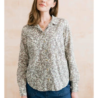 A woman wearing a long-sleeve button-up blouse with a dense small floral print in muted green, brown, and beige tones on an off-white base, paired with blue jeans against a soft beige background.