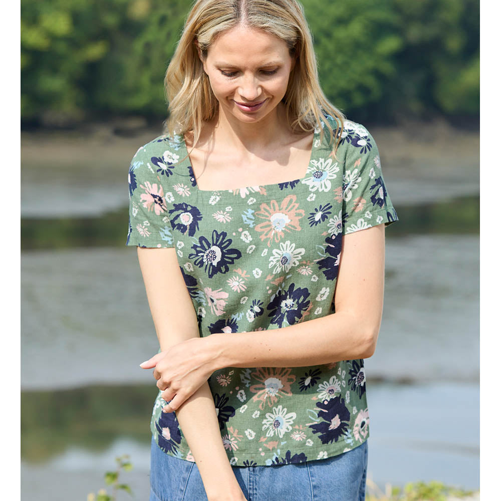 Blonde female wearing a khaki green floral top that has short sleeves and a squared neckline. The flowers are pale pink and navy blue