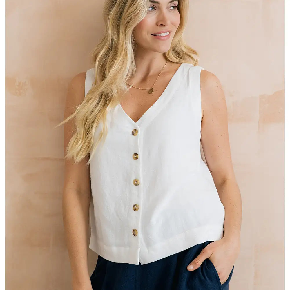 Woman wearing a white sleeveless blouse with gold buttons against a beige wall.