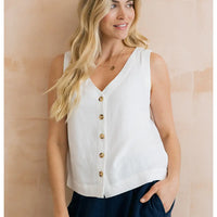 Woman wearing a white sleeveless blouse with gold buttons against a beige wall.