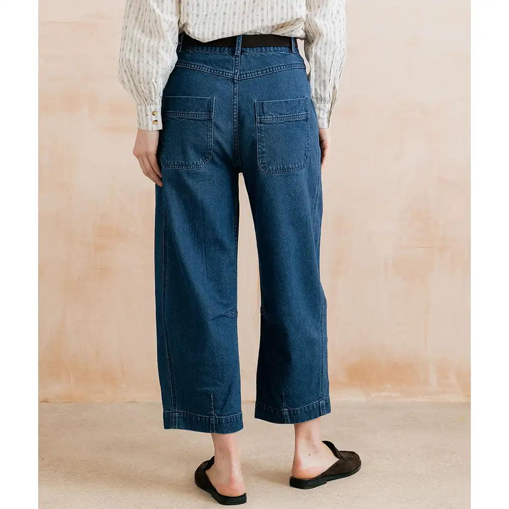 A person shown from the back wearing dark blue cropped wide-leg denim jeans, a cream striped long-sleeve blouse, and black flat mule sandals against a warm beige textured wall.