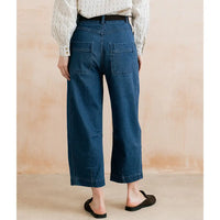 A person shown from the back wearing dark blue cropped wide-leg denim jeans, a cream striped long-sleeve blouse, and black flat mule sandals against a warm beige textured wall.