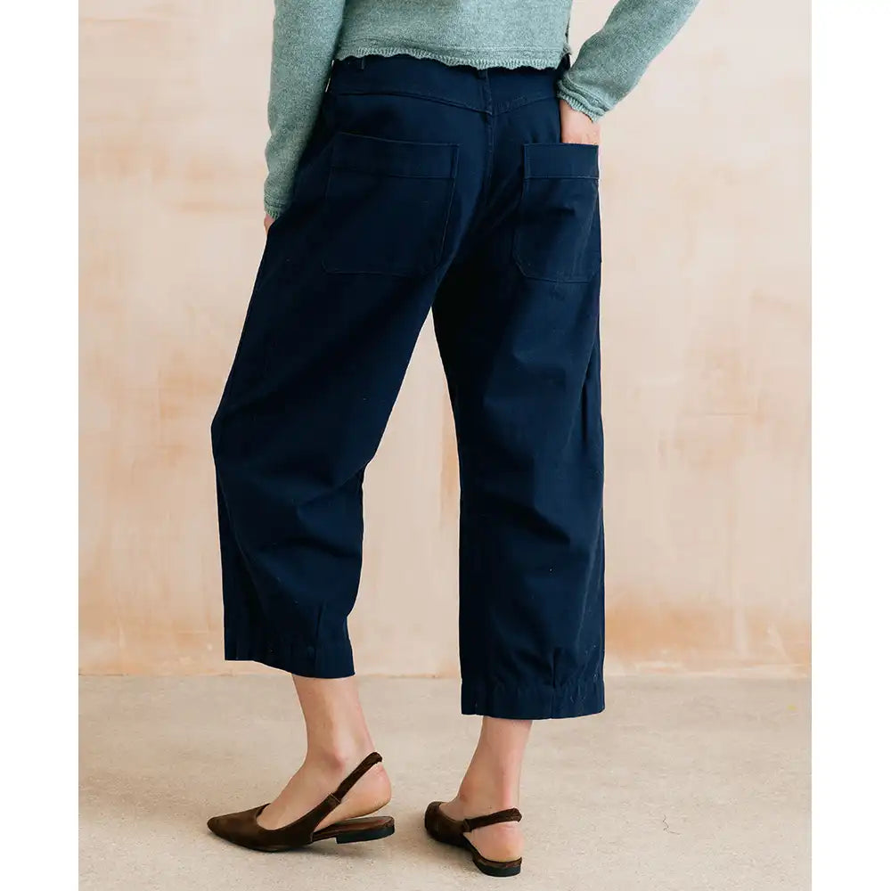 A person shown from the back wearing dark navy cropped pants with large patch pockets, a light green long-sleeve top, and dark brown slingback flats against a soft beige wall.