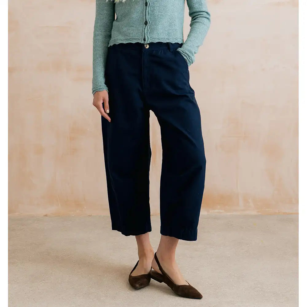 A model wearing dark navy cropped wide-leg trousers, a sage green cardigan, and brown slingback flats, standing against a textured beige wall.