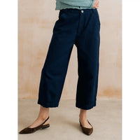 A person wearing dark navy cropped wide-leg pants with a light green top and brown slingback flats, standing against a warm beige textured wall.