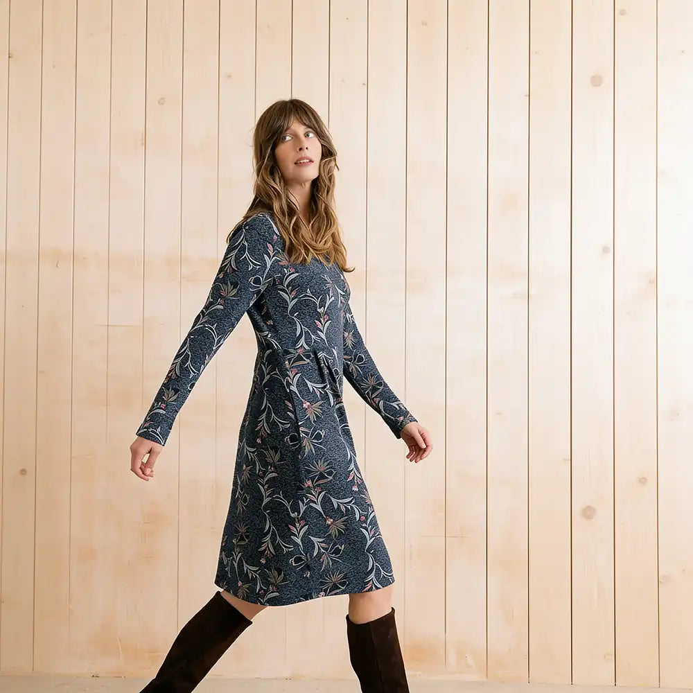 A woman wearing a long-sleeve navy blue dress with a white and beige floral pattern, paired with black knee-high boots, standing against a light wood-paneled wall.
