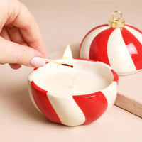 A hand is lighting a small white candle in a red-and-white striped bauble shaped holder, with a matching ornament-shaped candle lid and gold cap in the background.