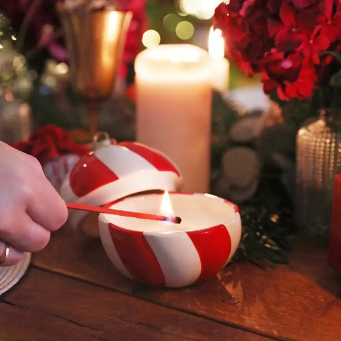 A hand holds a lit match to a small red-and-white striped candle bowl on a wooden table, with a lit pillar candle, red flowers, and holiday decor in the background.