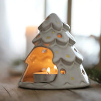 A ceramic Christmas tree-shaped candle holder with a small tea light candle inside, set on a wooden surface with a softly blurred indoor background.